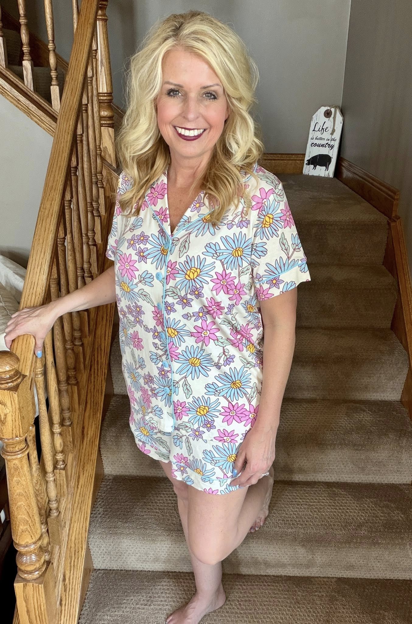 Woman wearing the LuLaRoe Candy PJ Set in a pastel daisy floral print featuring blue daisies, pink flowers, lavender blossoms, and yellow centers on an ivory background with a button-front pajama top and matching pocket shorts standing on a staircase