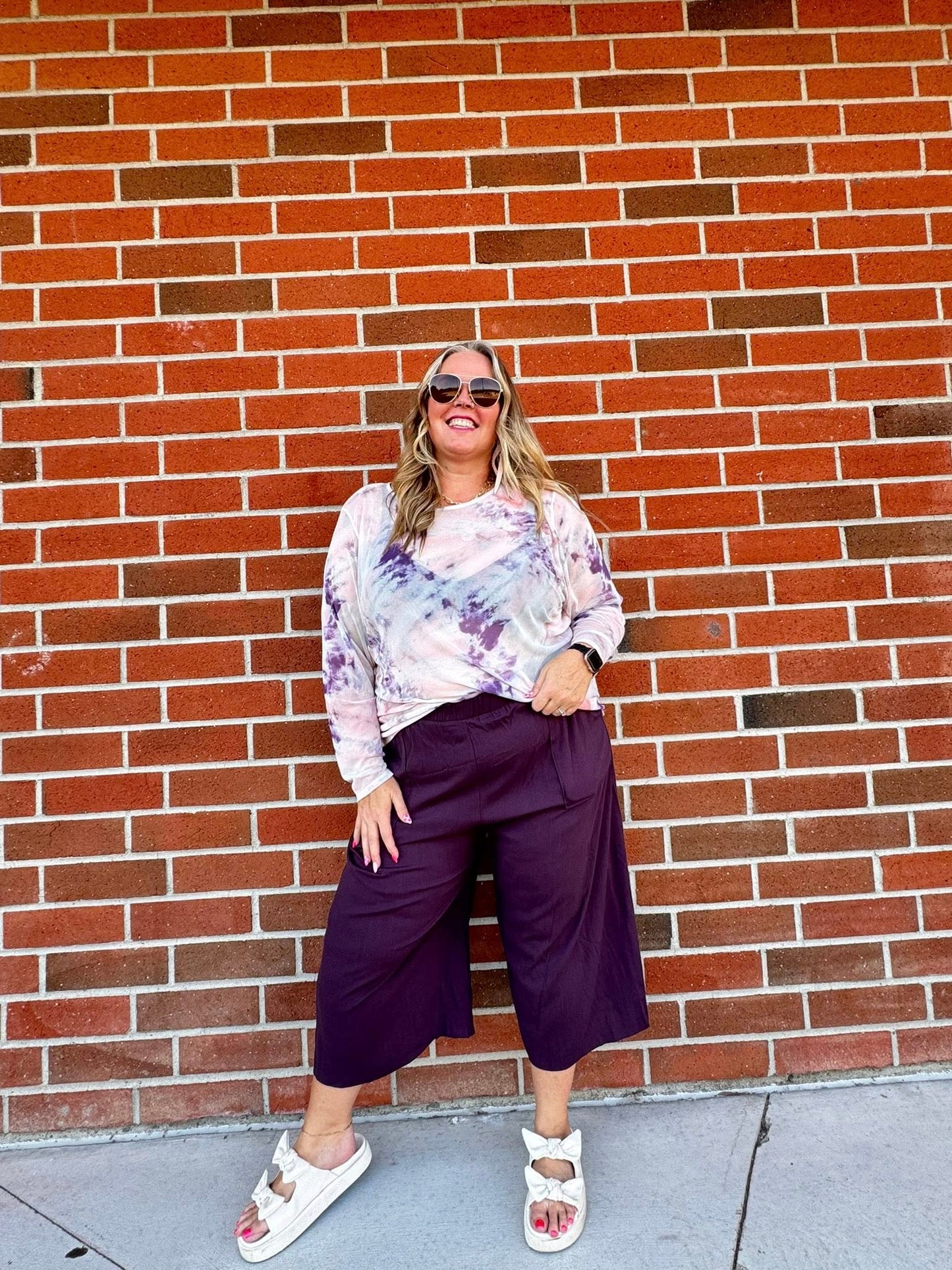 Woman wearing a tie-dye shirt and purple pants standing against a brick wall.
