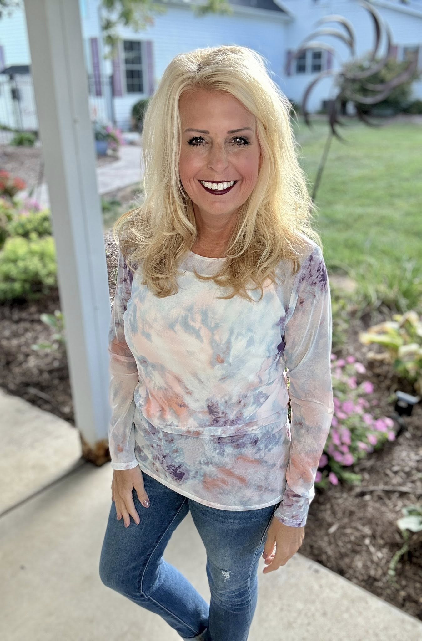Woman wearing a tie-dye shirt and jeans standing outdoors with a house and garden in the background.