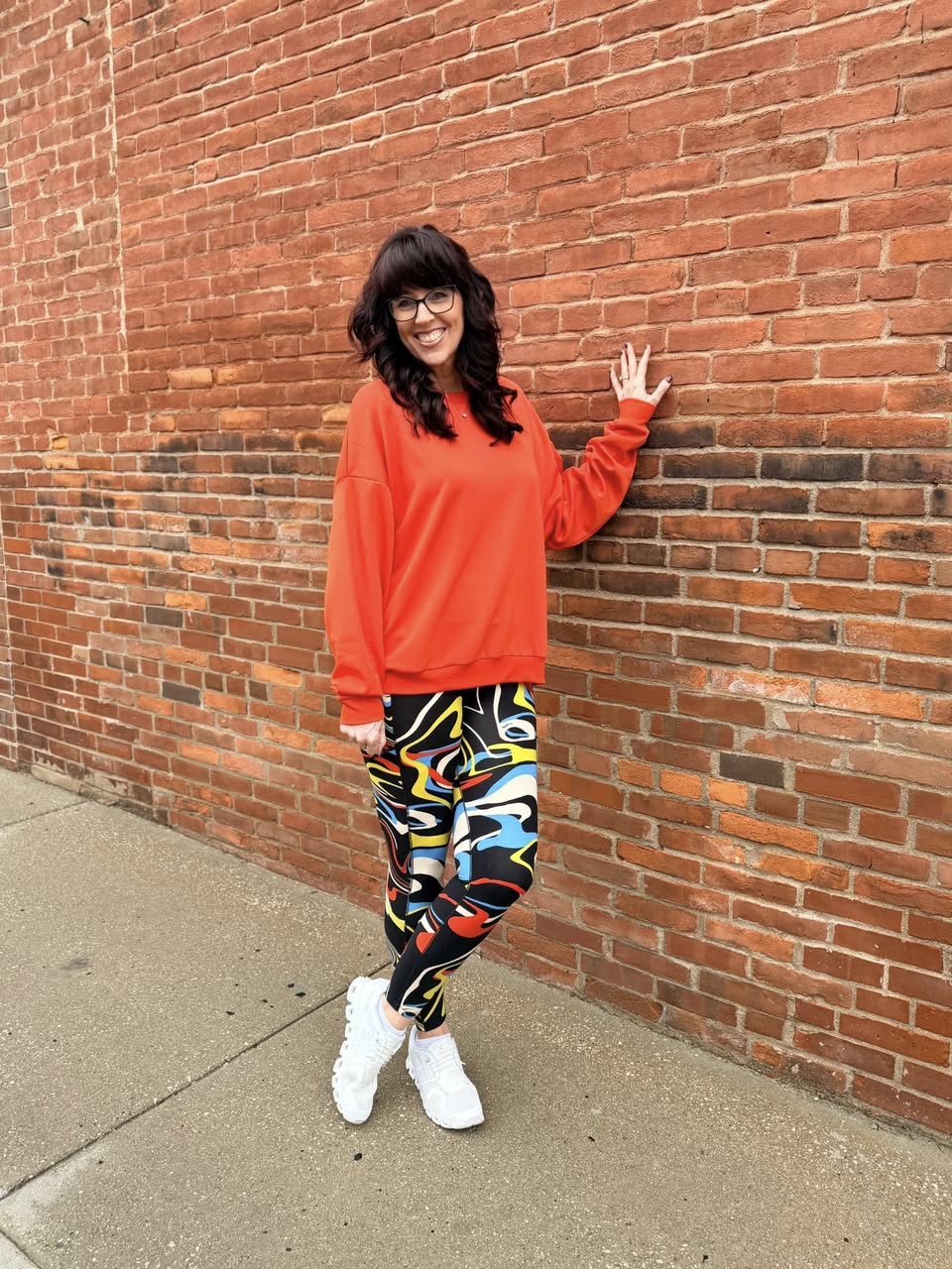 Person wearing a bright orange sweatshirt and colorful pants leaning against a brick wall.