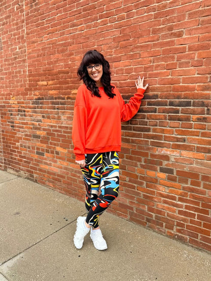 Person wearing a bright orange sweatshirt and colorful pants leaning against a brick wall.
