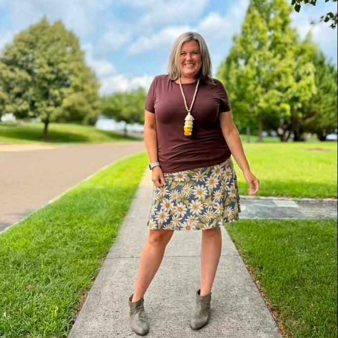 A woman stands on a paved path outdoors wearing a dark brown short-sleeve top, a yellow, white, and blue floral print skirt, and gray ankle boots. She is wearing a long gold pendant necklace.