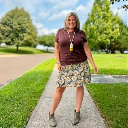A woman stands on a paved path outdoors wearing a dark brown short-sleeve top, a yellow, white, and blue floral print skirt, and gray ankle boots. She is wearing a long gold pendant necklace.