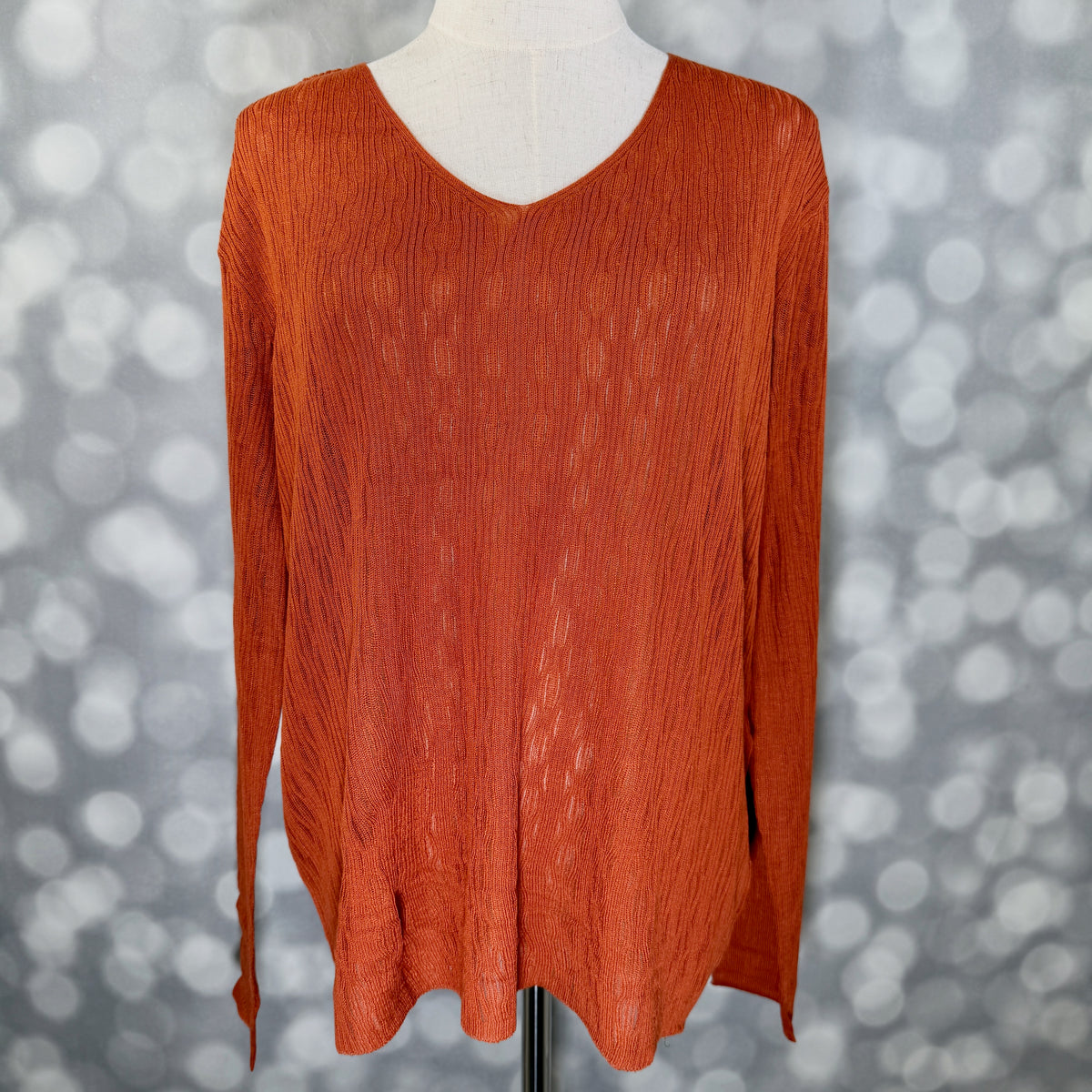 An orange long-sleeve V-neck knit top displayed on a mannequin torso against a gray bokeh background.