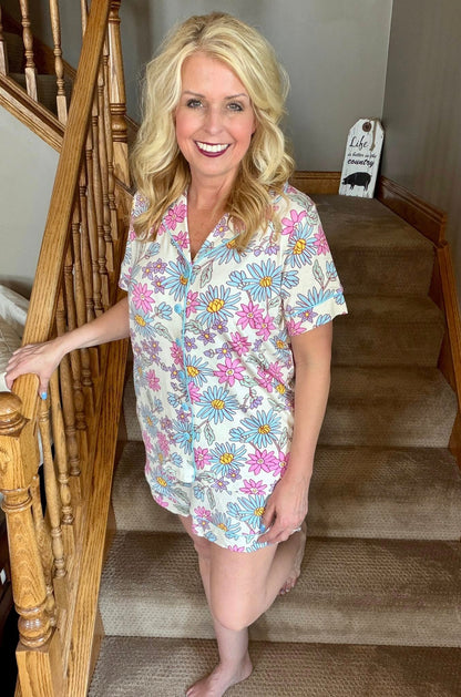 Woman wearing the LuLaRoe Candy PJ Set in a pastel daisy floral print featuring blue daisies, pink flowers, lavender blossoms, and yellow centers on an ivory background with a button-front pajama top and matching pocket shorts standing on a staircase