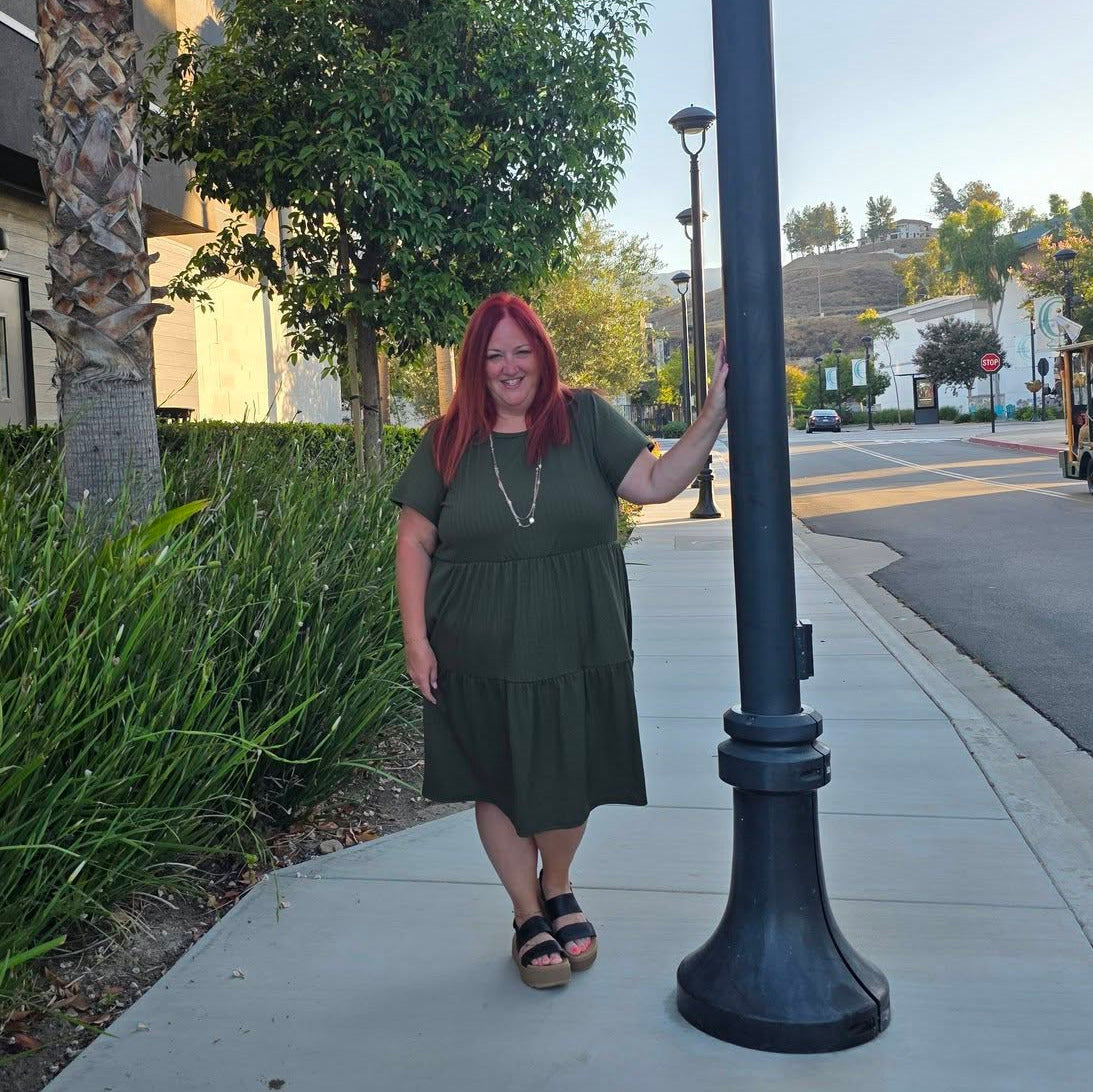 Woman standing on a sidewalk next to a palm tree and street lamp wearing a dark olive LuLaRoe Ariel dress.