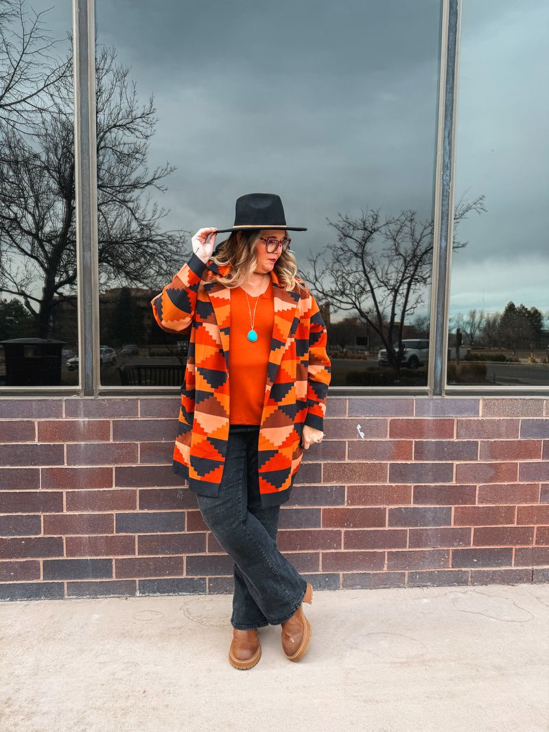 Model wearing an orange and black geometric Canyon Mosaic Laura lapel open cardigan with front pockets. standing in front of a brick wall.