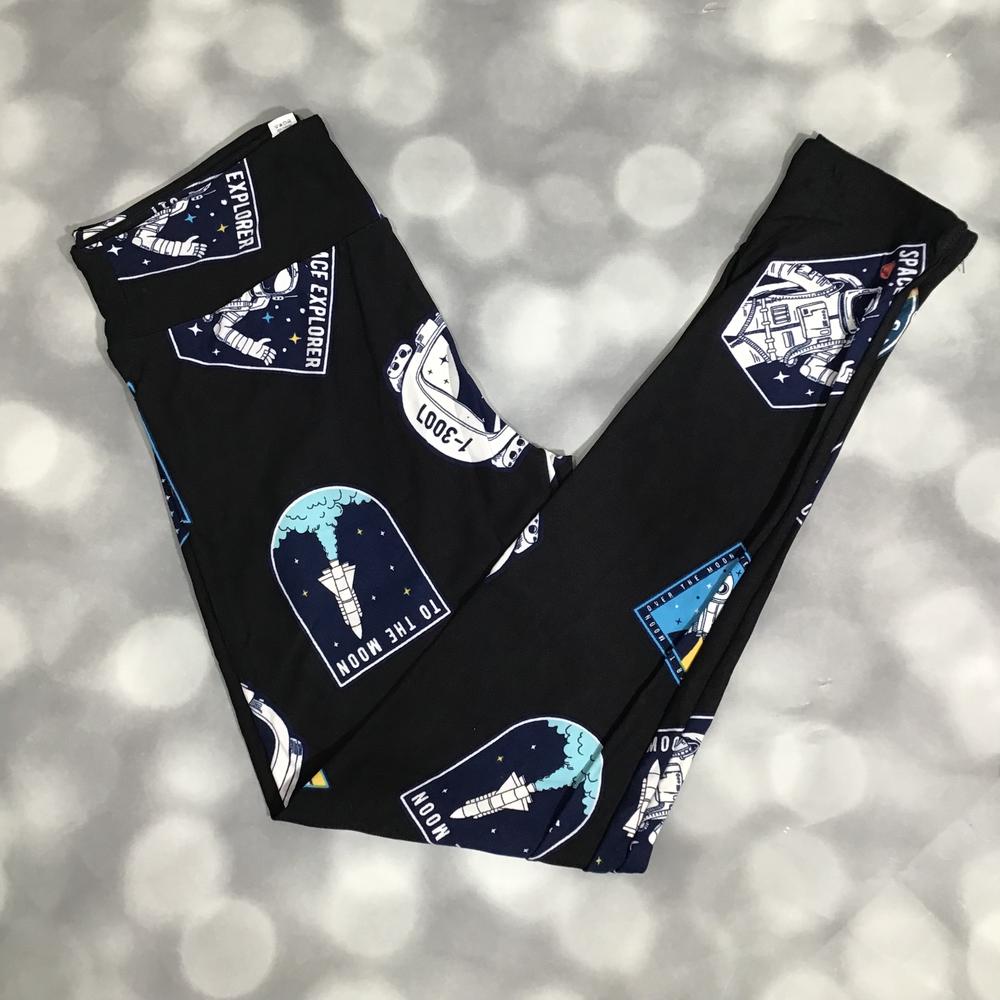 Flat lay of LuLaRoe leggings in black featuring novelty space patches, astronaut motifs, and rocket ship designs with a yoga waistband and ankle-length cut on a blurred background