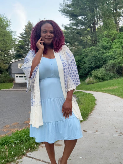Woman modeling the LuLaRoe Amanda Dress in Solid Baby Blue, featuring a soft pastel blue sleeveless tiered midi silhouette with a relaxed feminine fit with a lightweight patterned kimono standing on a sidewalk with greenery in the background.