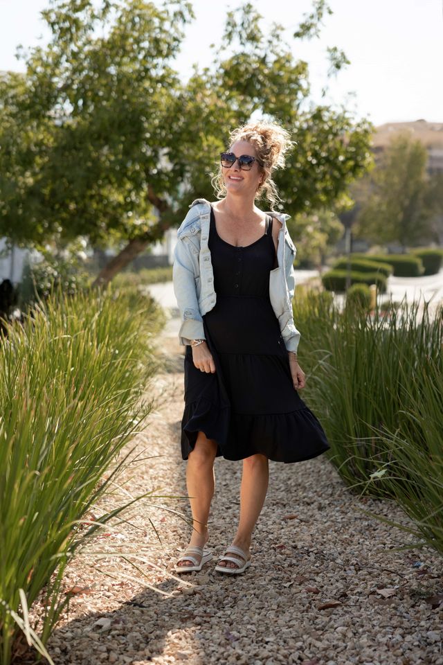 Woman modeling the LuLaRoe Amanda Dress in Solid Black, featuring a sleeveless black tiered midi silhouette with a relaxed flowy fit styled with a light denim jacket and neutral sandals outdoors in natural sunlight.