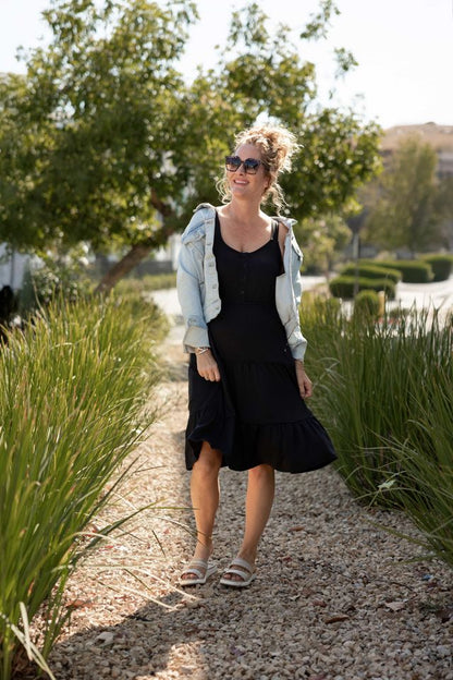 Woman modeling the LuLaRoe Amanda Dress in Solid Black, featuring a sleeveless black tiered midi silhouette with a relaxed flowy fit styled with a light denim jacket and neutral sandals outdoors in natural sunlight.