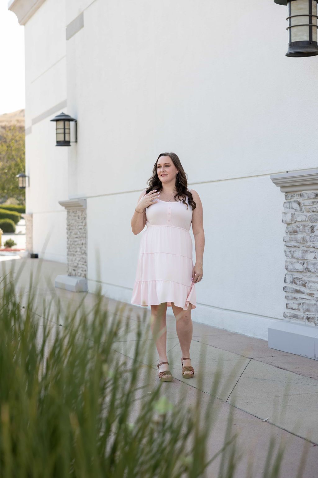 Woman modeling the LuLaRoe Amanda Dress in Solid Blush Taupe, featuring a soft blush beige sleeveless tiered midi silhouette with a relaxed feminine fit photographed outdoors in natural sunlight near a modern neutral building.