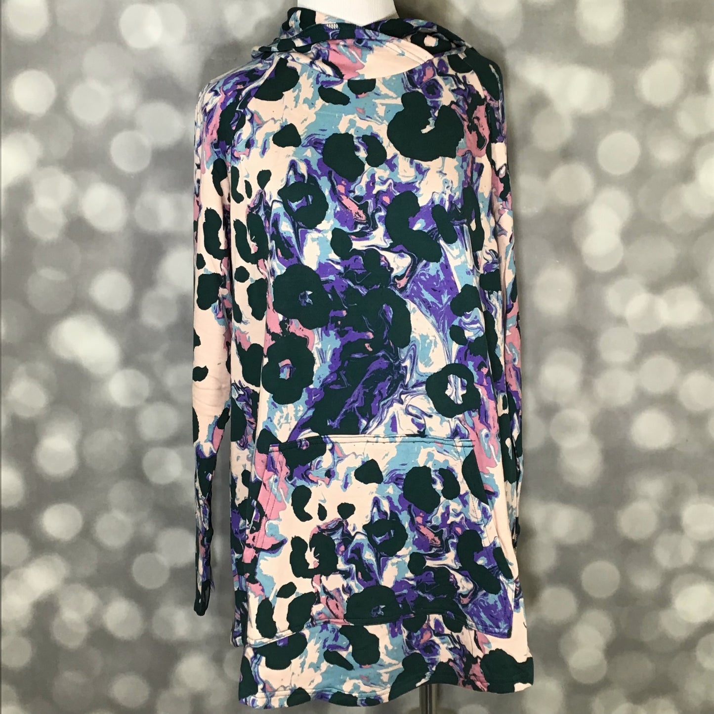 LuLaRoe Amber Hoodie in Amethyst Drift Abstract featuring a blush base with purple, blue, teal, and dark green abstract painterly print, showing long sleeves, hood, and kangaroo pocket with a front view on mannequin against a blurred background.