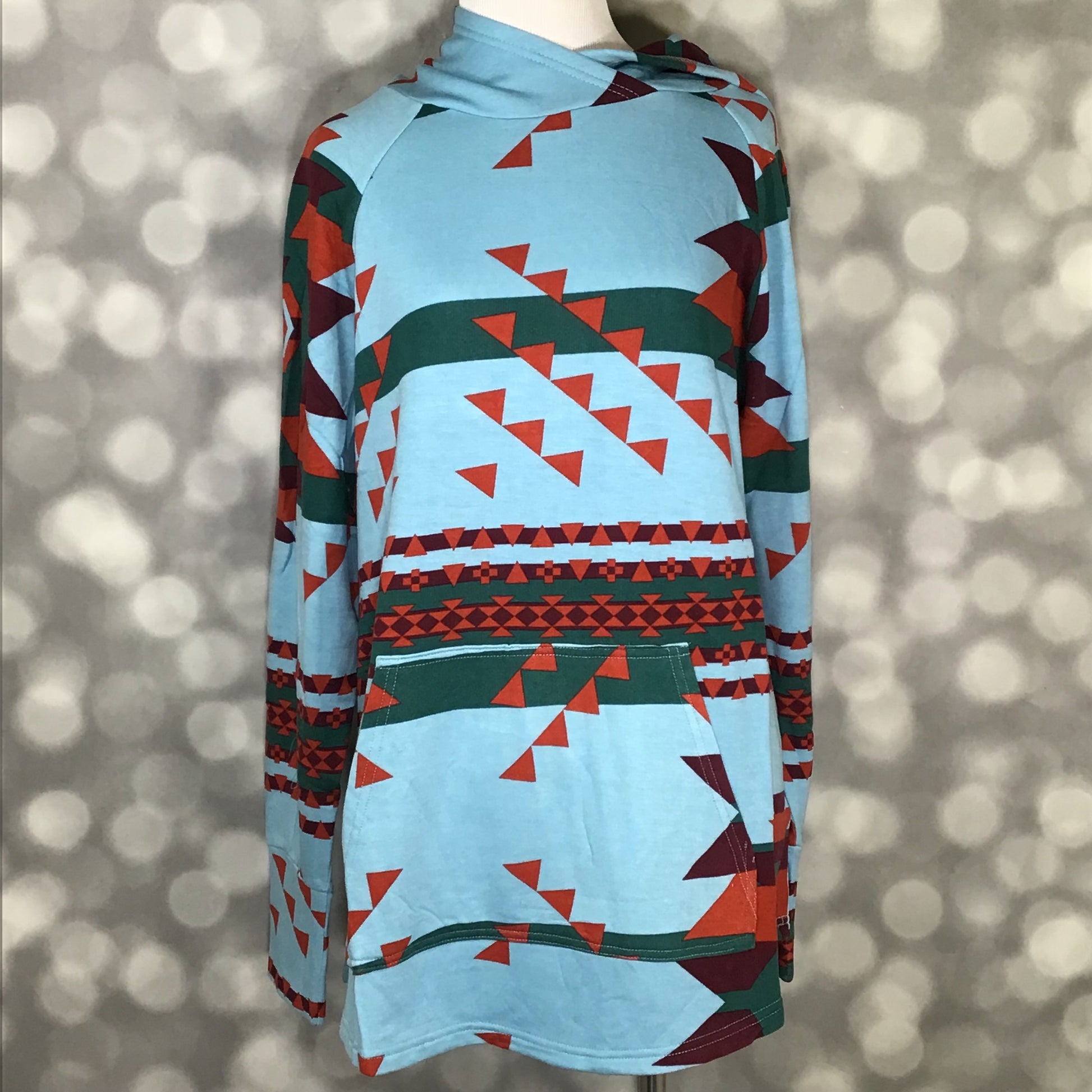 LuLaRoe Amber Hoodie in Teal Mesa Geometry with teal background, geometric triangle print, hood, and kangaroo pocket, front view on mannequin against a blurred background