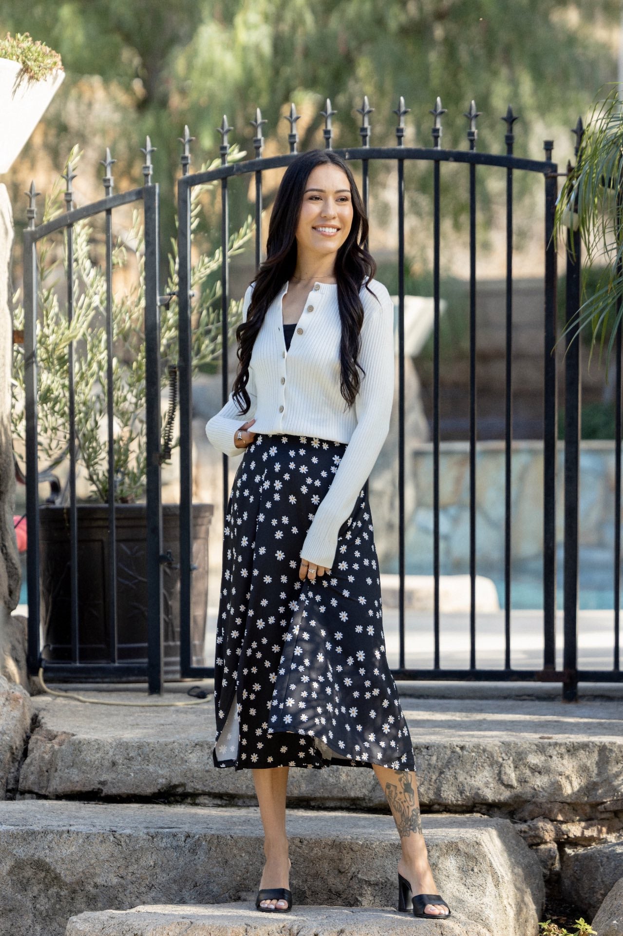 LuLaRoe Becky cardigan in solid soft ivory cream rib knit with wooden buttons and fitted silhouette on a woman in a white blouse and black polka dot skirt standing on steps with a black metal gate and trees in the background.