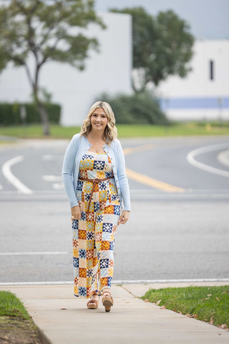 LuLaRoe Becky cardigan in solid soft sky blue rib knit with wooden buttons and fitted silhouette on a woman wearing a colorful patterned jumpsuit standing on a sidewalk with a road and trees in the background.
