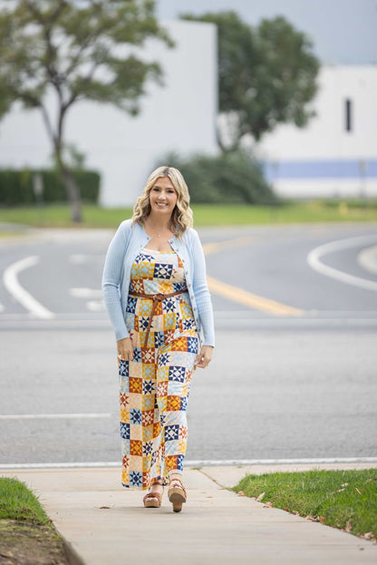 LuLaRoe Becky cardigan in solid soft sky blue rib knit with wooden buttons and fitted silhouette on a woman wearing a colorful patterned jumpsuit standing on a sidewalk with a road and trees in the background.