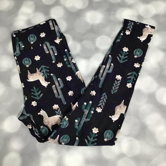 Flat lay of LuLaRoe leggings with whimsical white llamas, green cacti, and desert plants printed on a black background on a textured background