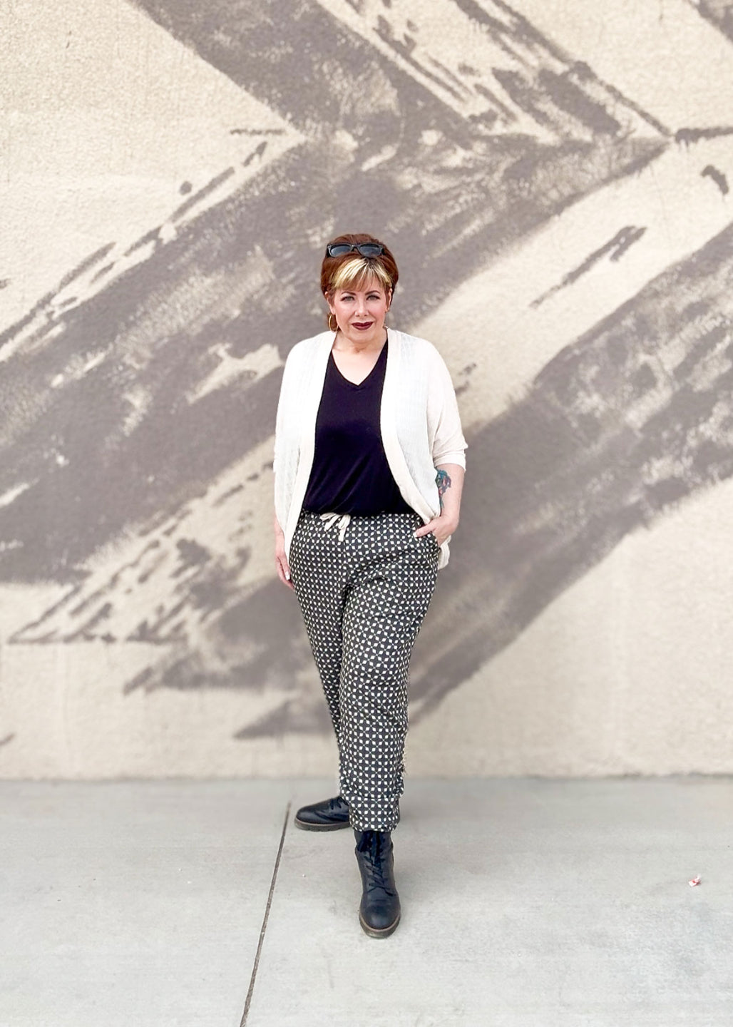 Woman standing in front of a textured wall wearing a white cardigan, black top, and patterned pants. LuLaRoe Boyfriend Joggers in a black and cream geometric tile print worn by a model outdoors. Relaxed-fit joggers with drawstring waist, cuffed ankles, and soft Ponte fabric.