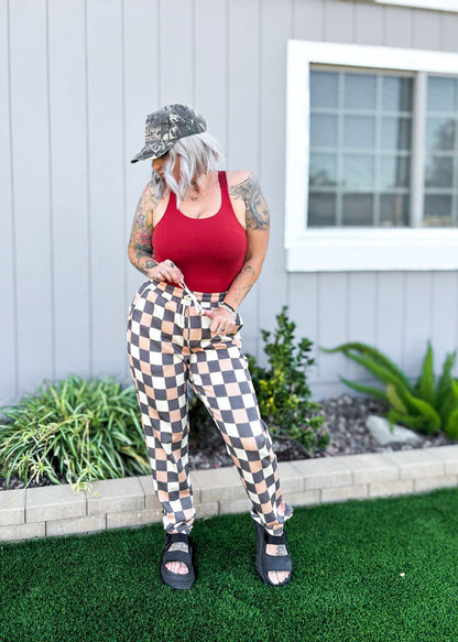 Person wearing a red tank top and checkered pants standing in front of a house. LuLaRoe Boyfriend Joggers in a tan and black checkerboard print worn outdoors by a model. Relaxed-fit joggers with drawstring waist, cuffed ankles, and soft Ponte fabric.