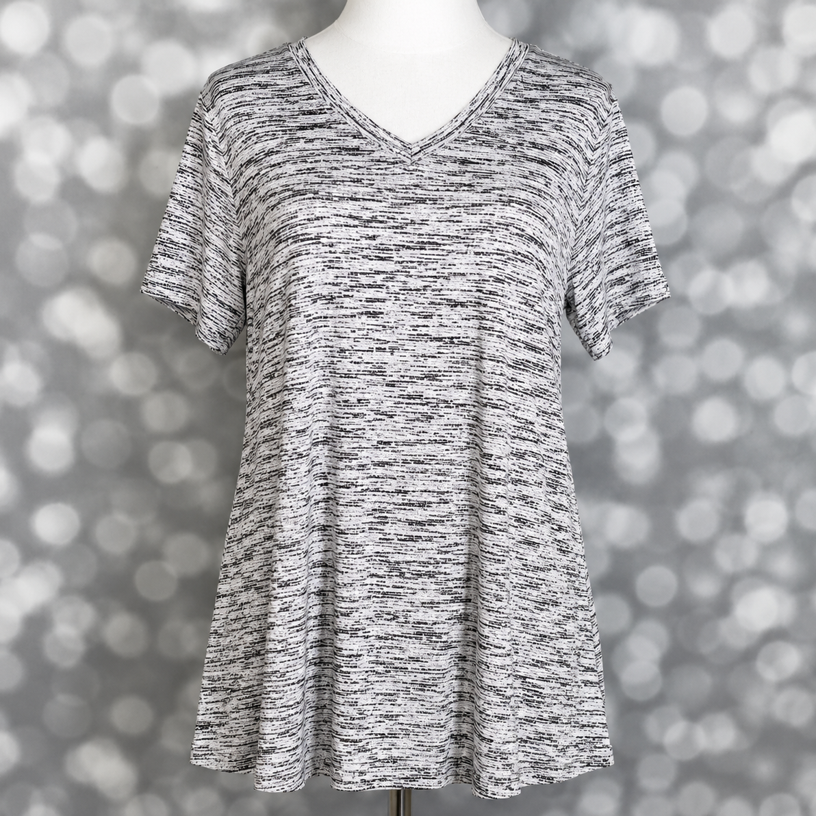 LuLaRoe Christy T relaxed swing top in black and white static heather print with subtle marled texture, v-neckline, short sleeves, and flowy silhouette displayed on mannequin against silver bokeh background.