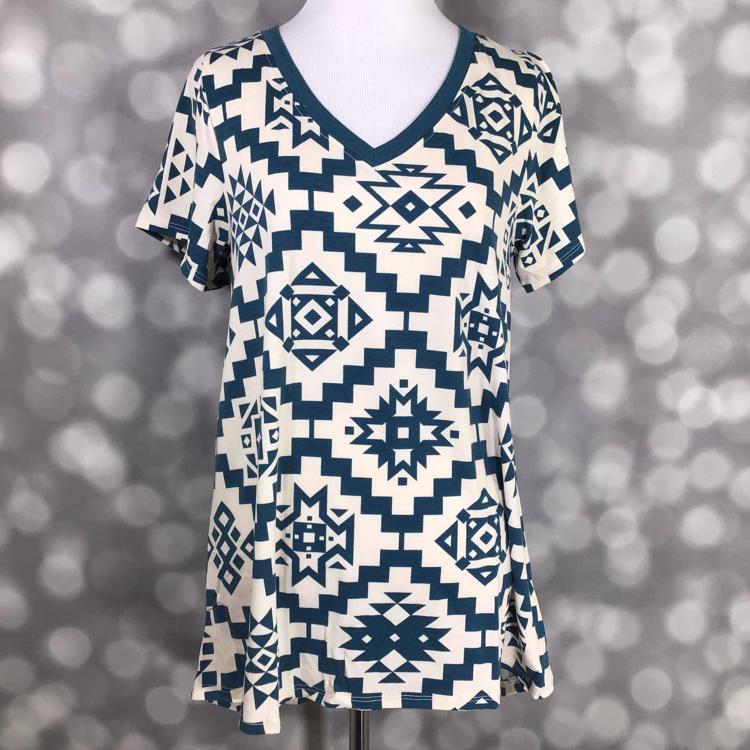 LuLaRoe Christy T featuring muted teal Southwestern geometric tribal patterns on an ivory background with a relaxed V-neck swing silhouette displayed on a mannequin against a silver bokeh backdrop.