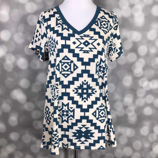 LuLaRoe Christy T featuring muted teal Southwestern geometric tribal patterns on an ivory background with a relaxed V-neck swing silhouette displayed on a mannequin against a silver bokeh backdrop.