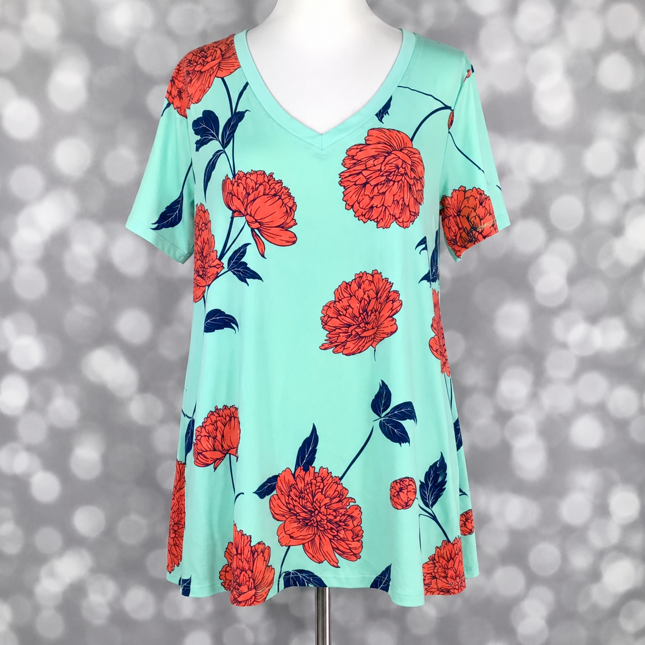 LuLaRoe Christy T swing tee in mint aqua featuring oversized coral floral sketch blooms with navy leaves and stems, short sleeves, v-neckline, and relaxed flowy silhouette displayed on mannequin against silver bokeh background.