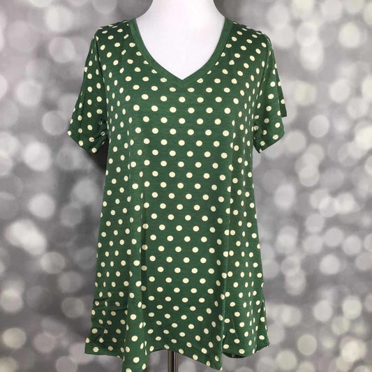 LuLaRoe Christy Tee featuring an olive green background with cream polka dots, displayed on a mannequin with a relaxed swing silhouette, short sleeves, and v-neckline against a gray bokeh background. Vintage-inspired polka dot boutique top with an easy everyday fit and soft casual styling.