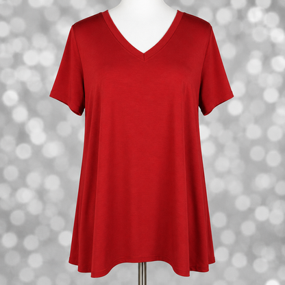LuLaRoe Christy Tee in solid true red displayed on a mannequin with a relaxed swing silhouette, short sleeves, and v-neckline against a soft gray bokeh background. Bright red boutique tunic top with an easy flowy fit perfect for casual everyday styling and layering.