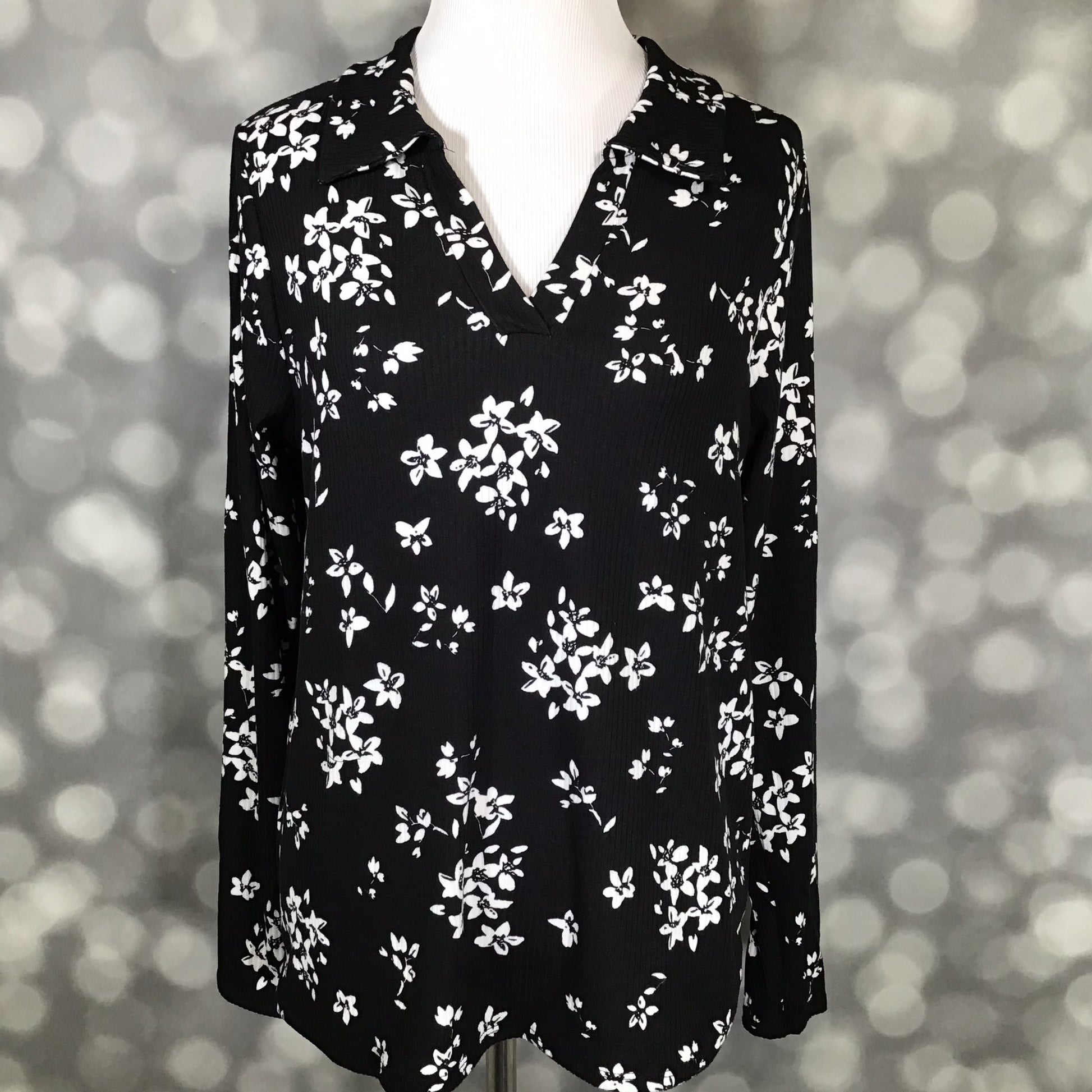 LuLaRoe Courtney Long Sleeve Polo Top in black with white scattered floral print, displayed on a mannequin against a soft light-bokeh background. Features a classic polo collar, V-notch neckline, long sleeves, and a comfortable draped fit in soft 4x2 Rib Brushed fabric.