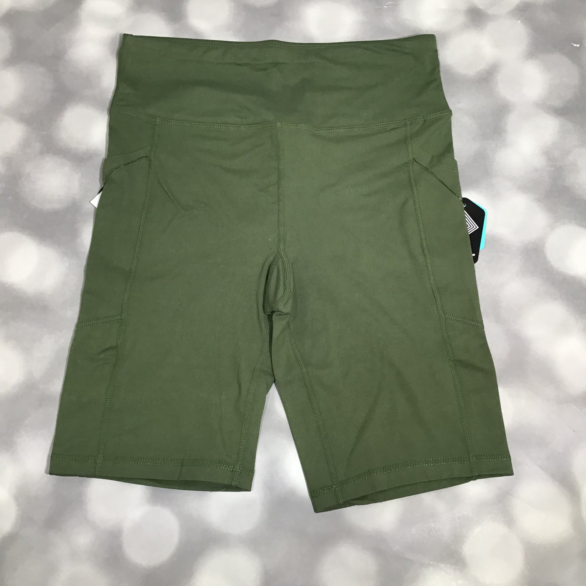 LuLaRoe Driven Shorts in solid olive green, displayed flat on a gray bokeh background. The shorts feature a high-rise waistband, side pockets, and mid-thigh length in smooth Lotus Jersey fabric.