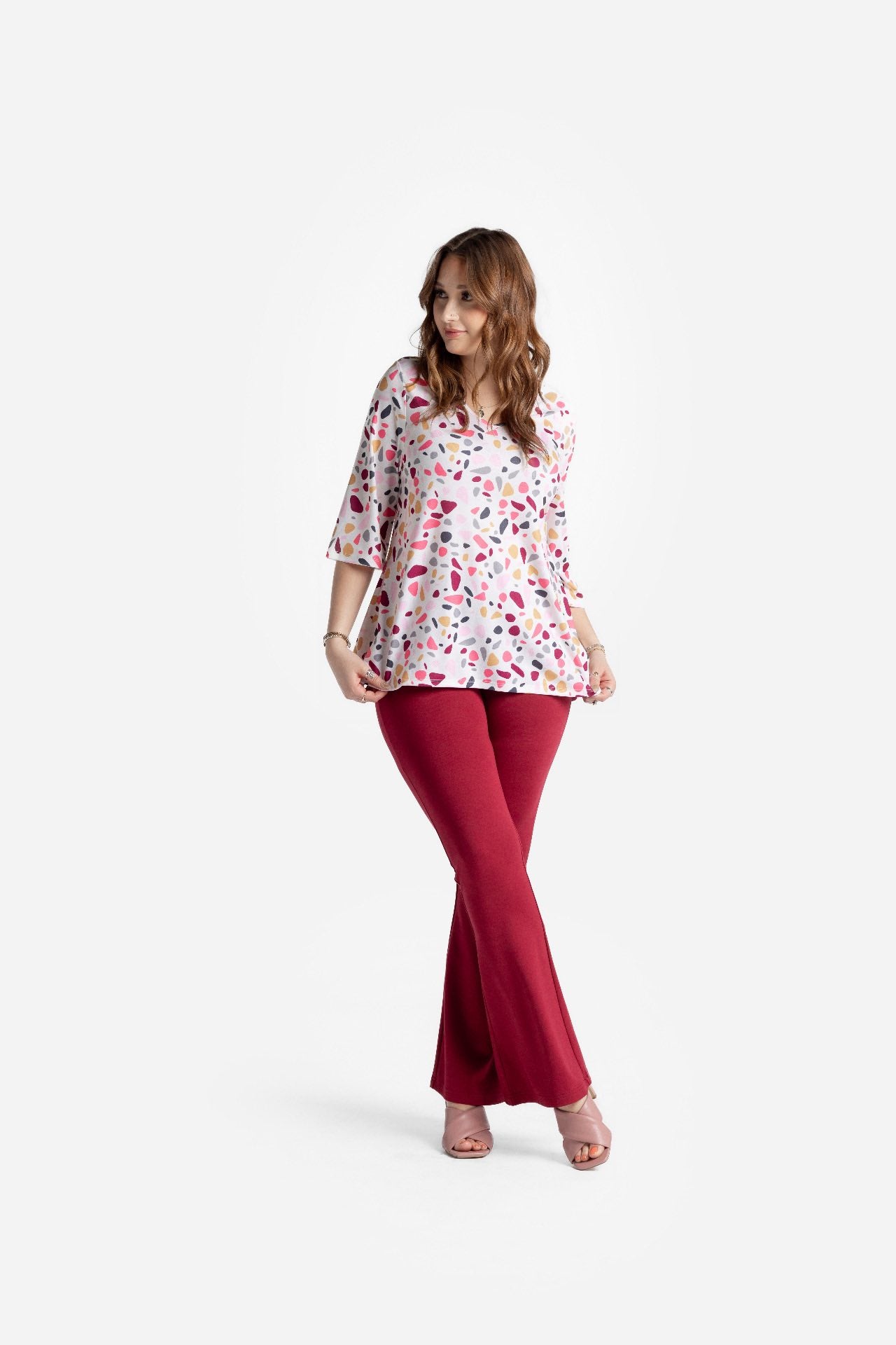 LuLaRoe Eden top in blush pink with multicolor terrazzo style abstract petal print, featuring three quarter sleeves and a flowy high low hem, worn with red pants in a studio setting on a white background