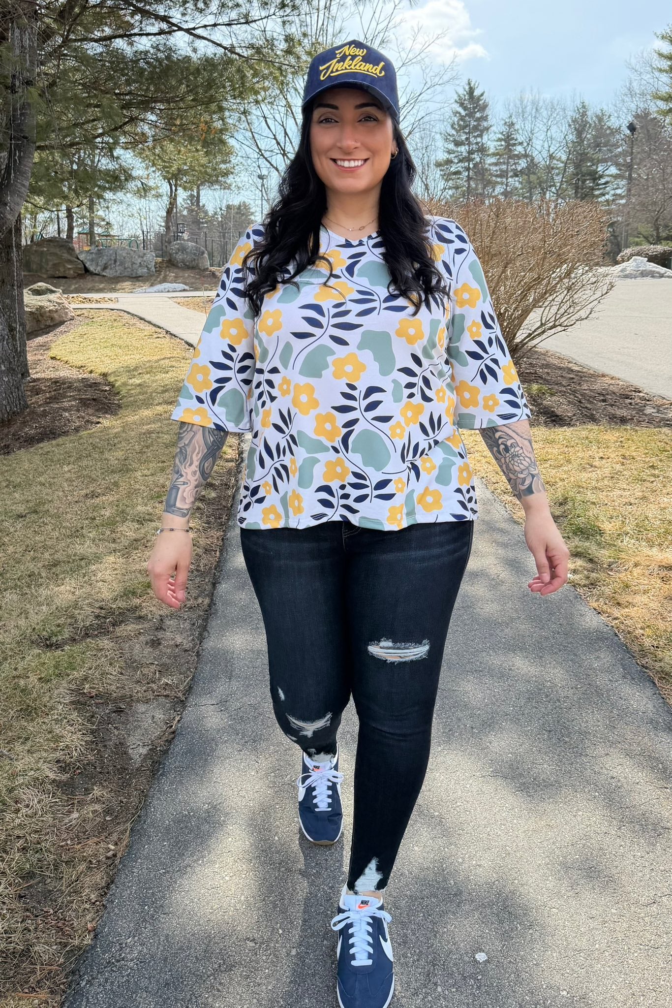 Woman walking outdoors wearing LuLaRoe Eden top with abstract floral print featuring yellow flowers, sage green shapes, and navy leaves on a light background, styled with dark jeans and sneakers.