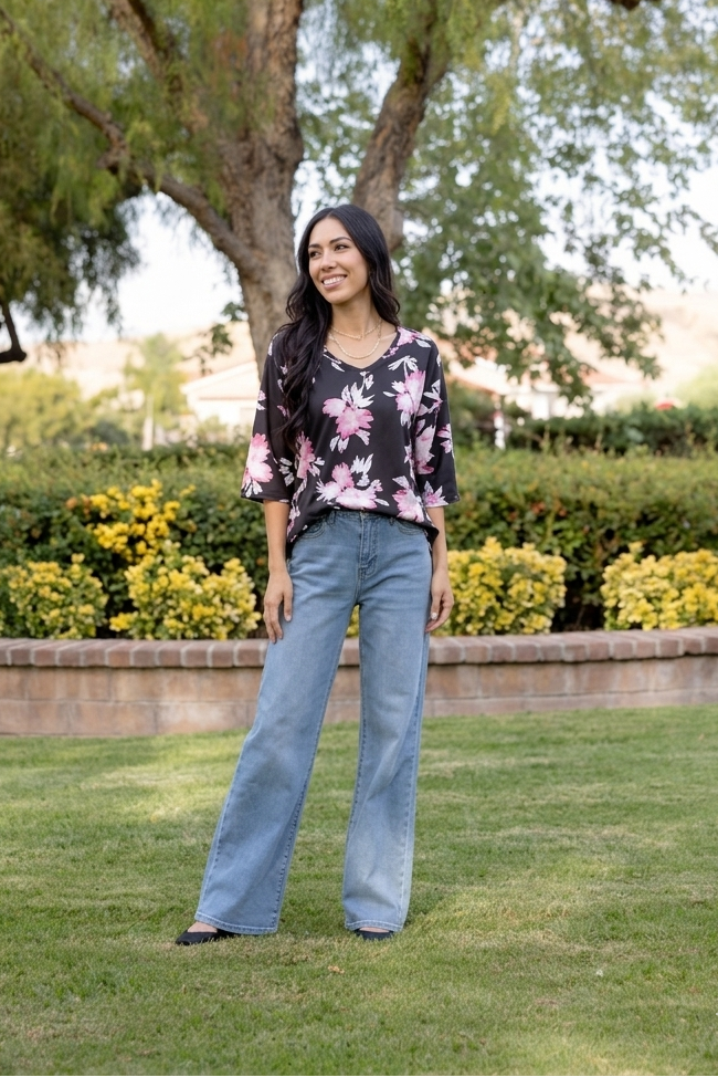Woman standing in a park with trees and bushes in the background wearing LuLaRoe Eden top with black background and blush pink floral print, featuring three quarter sleeves and a flowy relaxed fit, styled with light wash wide leg jeans .