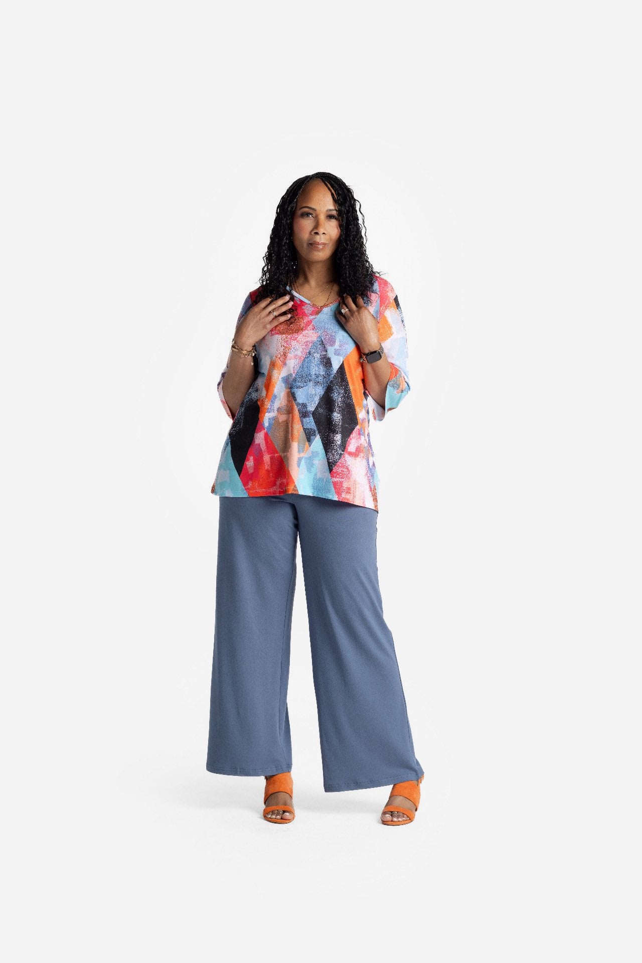 LuLaRoe Eden top featuring a colorful abstract geometric print in coral, teal, blue, and orange tones with three quarter sleeves and a flowy relaxed fit, styled with blue wide leg pants and orange heels on model on a white background