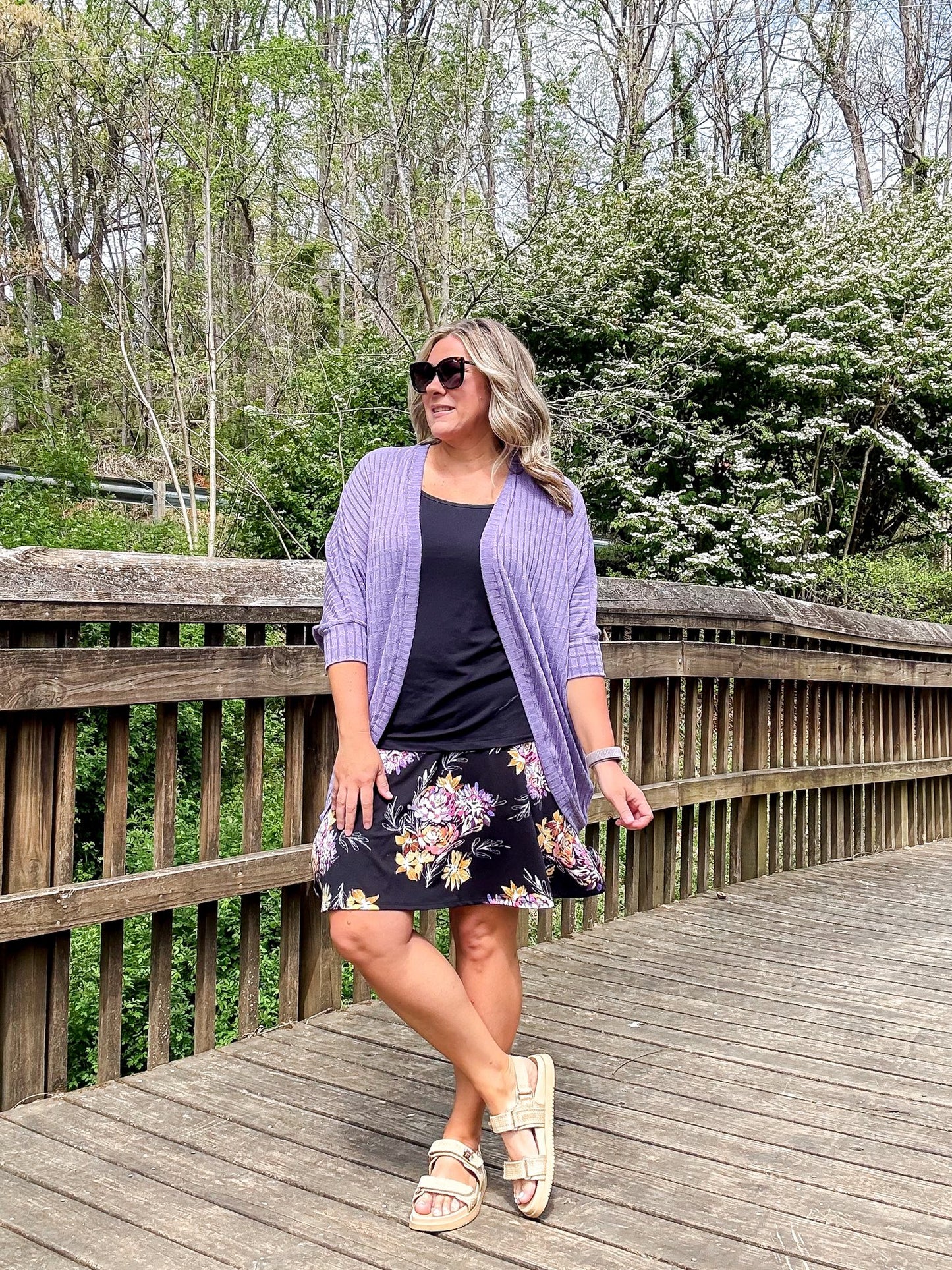 Woman wearing LuLaRoe Eileen skort in black with soft floral print in pink and cream tones, featuring a flared silhouette with built-in shorts, styled with a black top and lavender cardigan standing on a wooden bridge with trees in the background