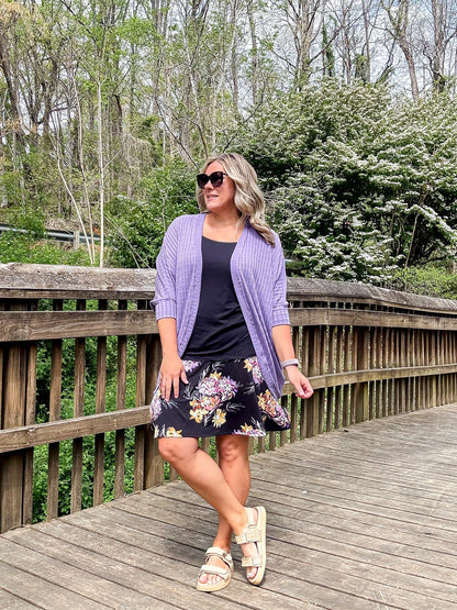 Woman wearing LuLaRoe Eileen skort in black with soft floral print in pink and cream tones, featuring a flared silhouette with built-in shorts, styled with a black top and lavender cardigan standing on a wooden bridge with trees in the background