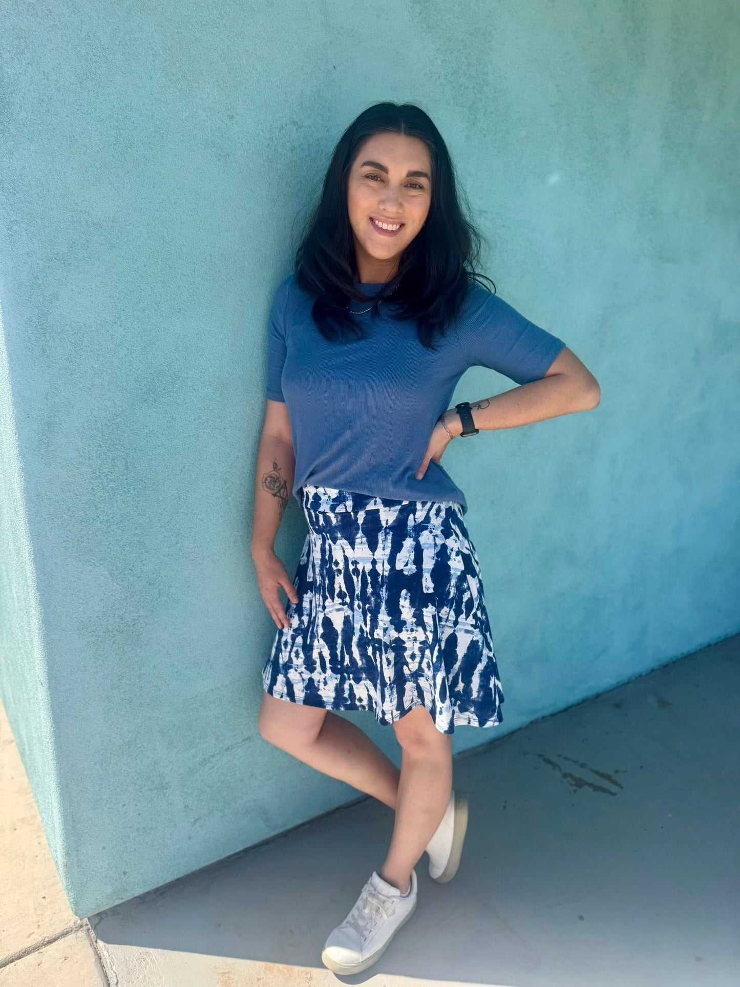 Woman in LuLaRoe Eileen skort in blue and white tie-dye shibori print with flared silhouette and built-in shorts, styled with a blue top and white sneakers standing against a turquoise wall.
