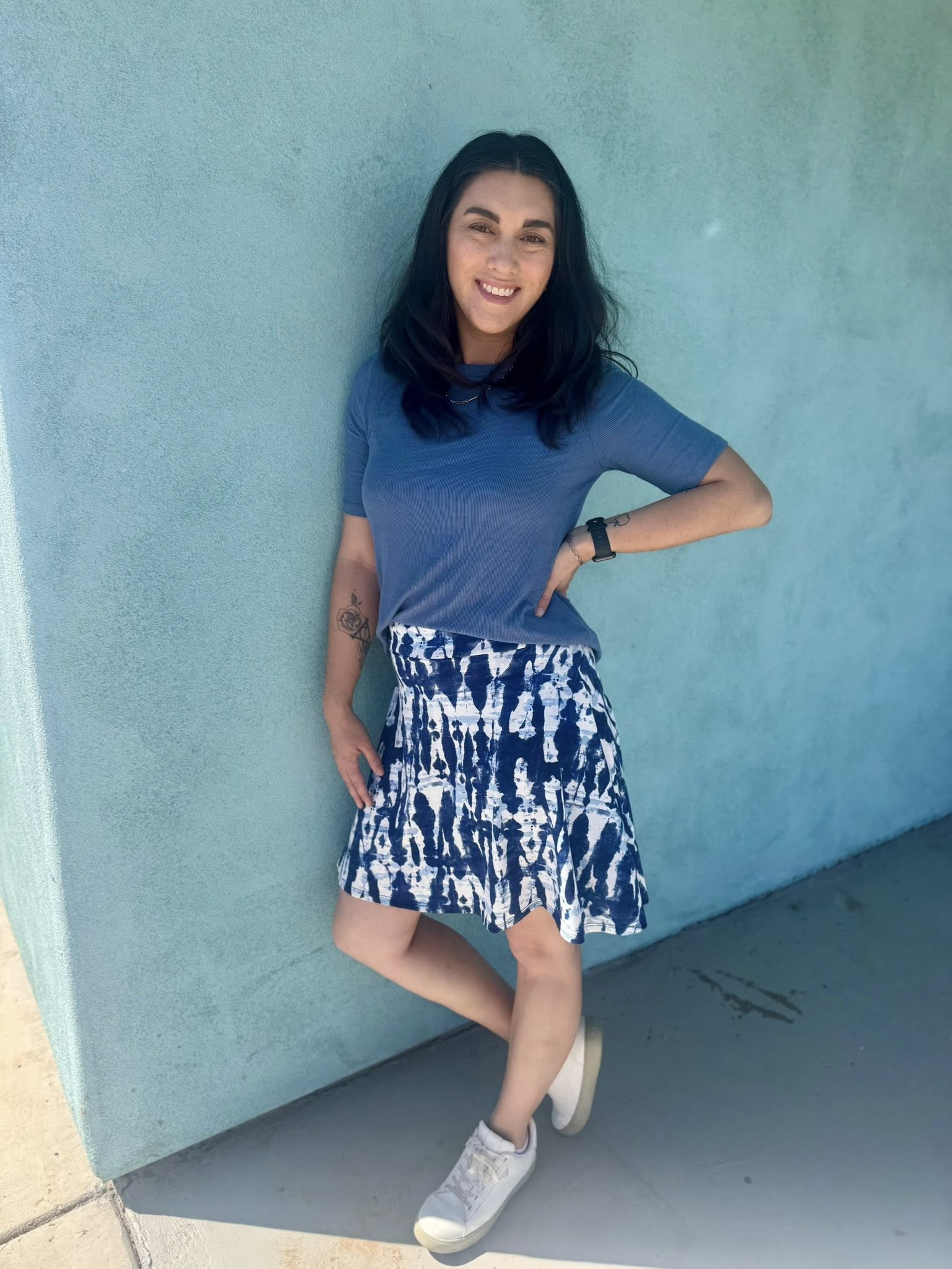 Woman in LuLaRoe Eileen skort in blue and white tie-dye shibori print with flared silhouette and built-in shorts, styled with a blue top and white sneakers standing against a turquoise wall.