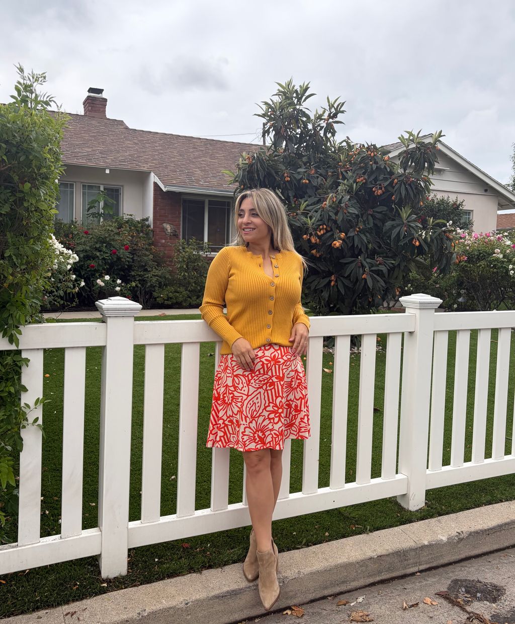 Woman wearing LuLaRoe Eileen skort in ivory with coral red floral sketch print, featuring a flared silhouette with built-in shorts, styled with a mustard cardigan standing in front of a white picket fence with a house in the background.