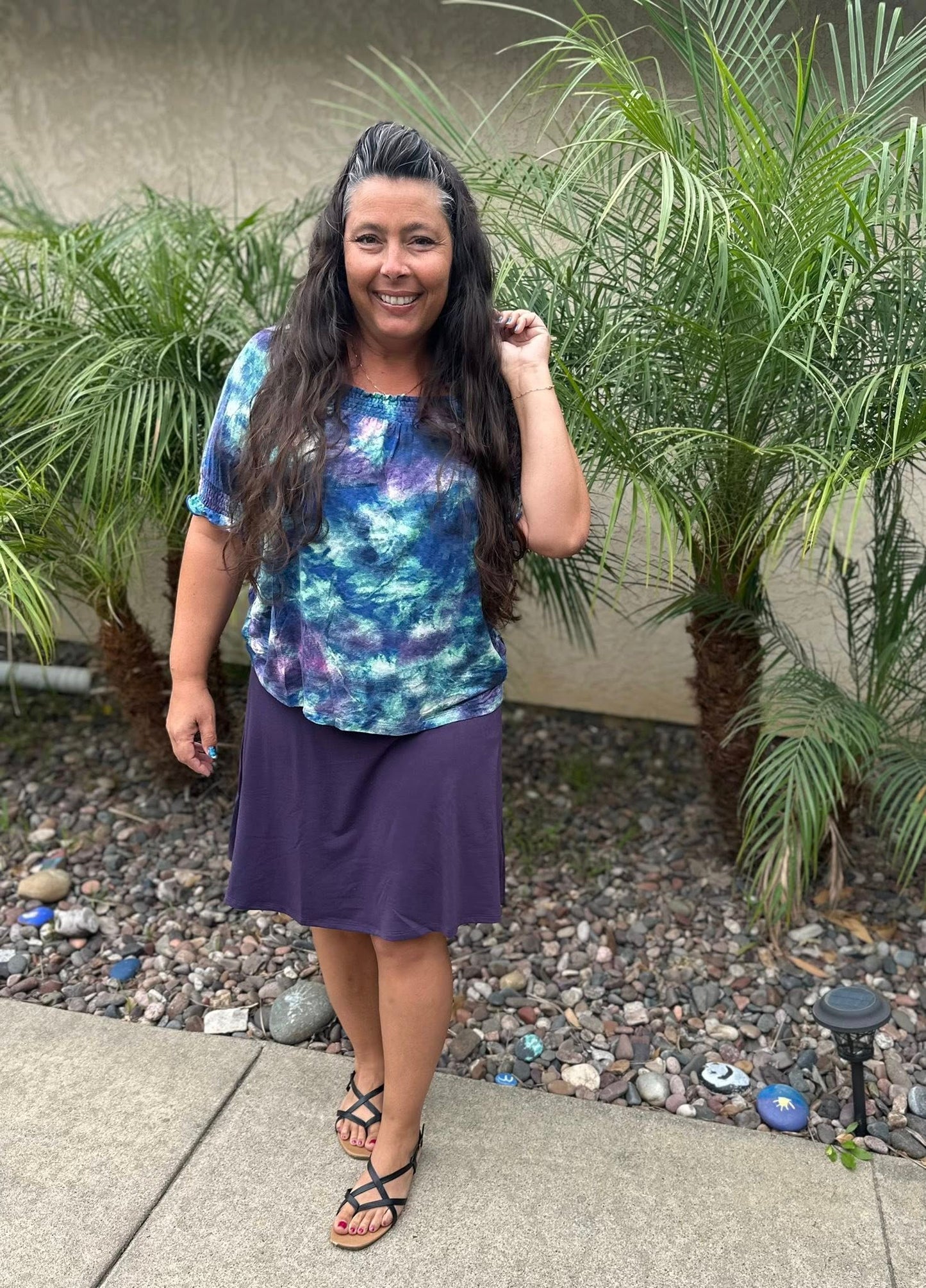 Woman wearing LuLaRoe Eileen skort in solid deep plum purple with flared A-line silhouette and built-in shorts, styled with a blue and purple tie-dye top standing outdoors with plants in the background