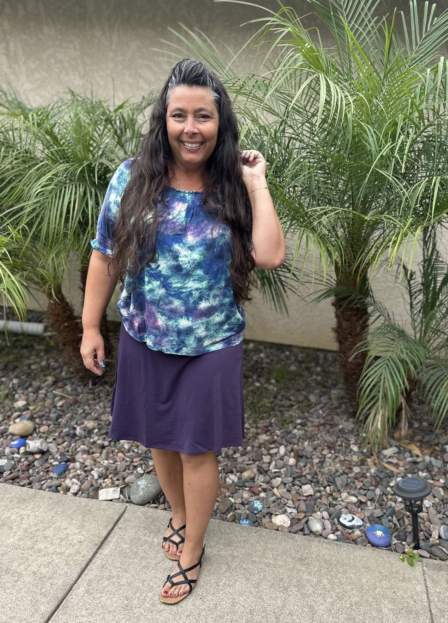 Woman wearing LuLaRoe Eileen skort in solid deep plum purple with flared A-line silhouette and built-in shorts, styled with a blue and purple tie-dye top standing outdoors with plants in the background