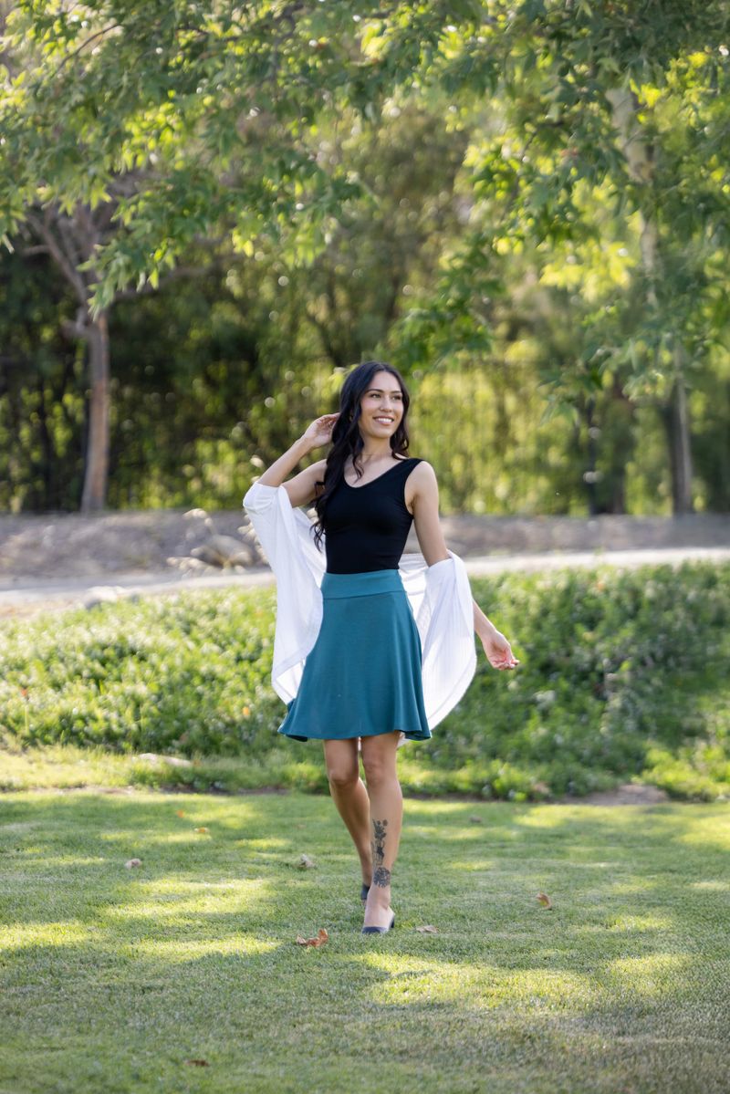 Woman walking outdoors in a park with trees and grass in the background wearing LuLaRoe Eileen skort in solid deep teal color with flared silhouette and built-in shorts, styled with a black tank and lightweight white wrap.