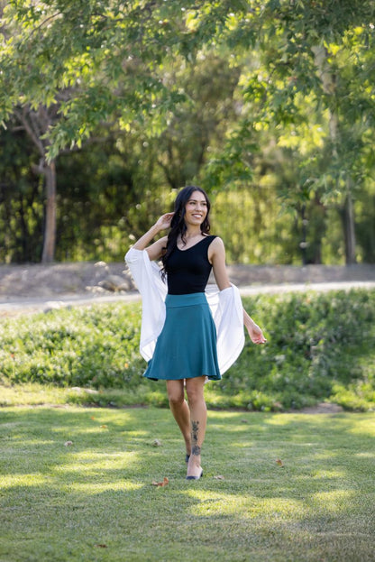 Woman walking outdoors in a park with trees and grass in the background wearing LuLaRoe Eileen skort in solid deep teal color with flared silhouette and built-in shorts, styled with a black tank and lightweight white wrap.