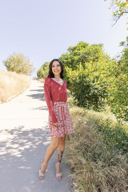 Woman wearing LuLaRoe Eileen skort in rustic red with ivory geometric boho mosaic print, featuring a flared skirt silhouette with built-in shorts, styled outdoors with a long sleeve top standing on a path surrounded by greenery