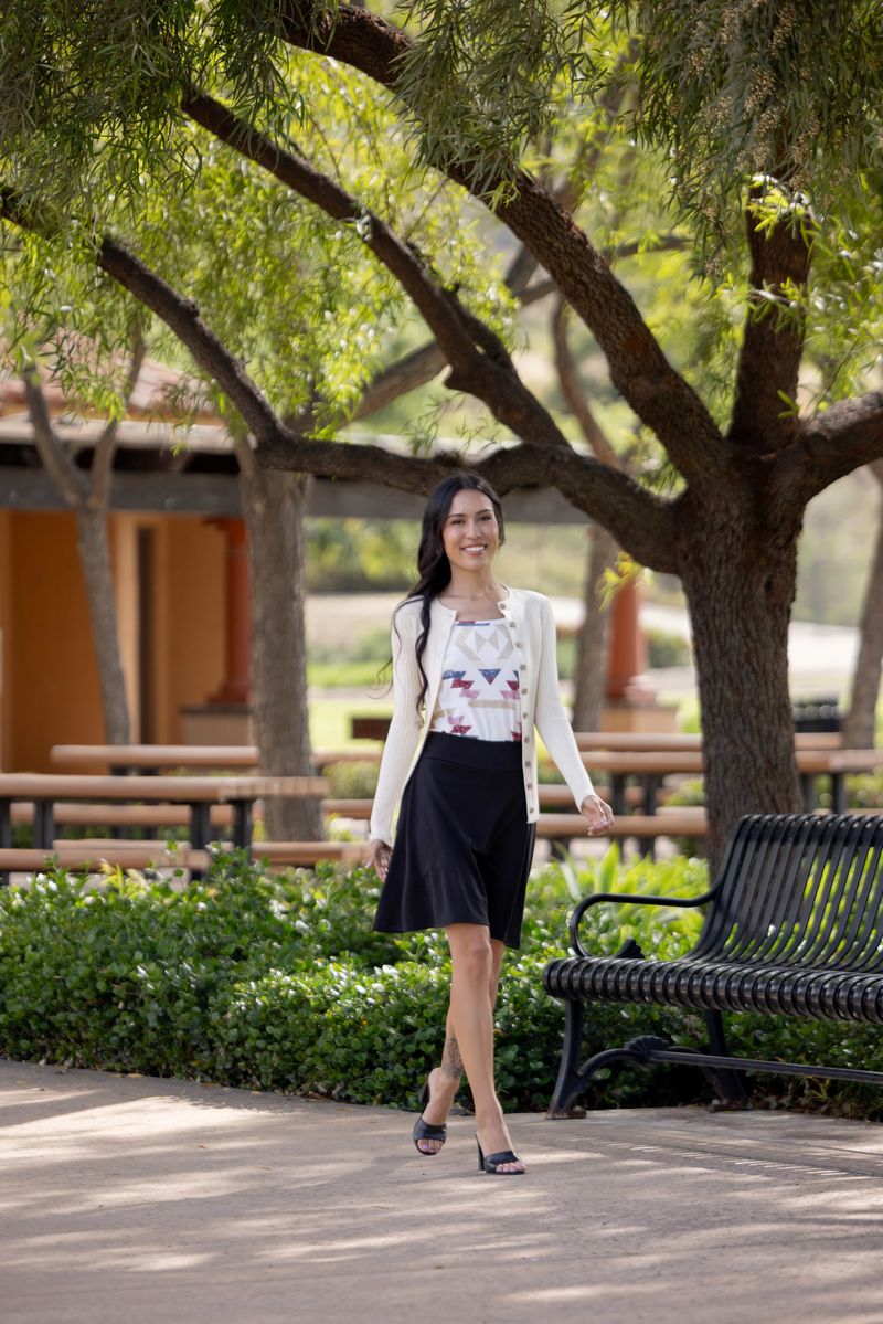 Woman wearing LuLaRoe Eileen skort in solid black featuring a smooth pull-on waistband with built-in shorts, worn styled with a printed top and cardigan outdoors in a park-like setting with trees and benches.