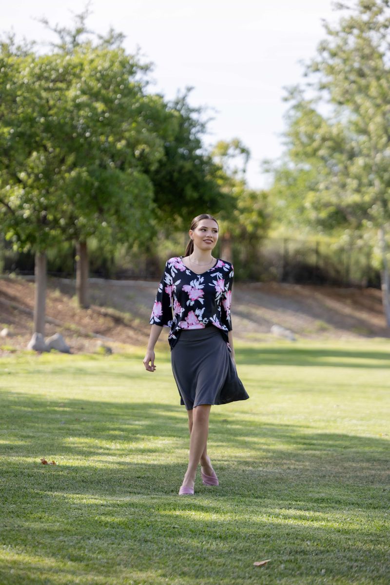Woman walking on grass with trees in the background LuLaRoe Eileen skort in solid charcoal gray with a pull-on waistband and built-in shorts, styled with a floral top outdoors on a grassy park setting