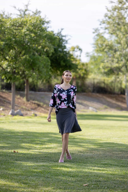 Woman walking on grass with trees in the background LuLaRoe Eileen skort in solid charcoal gray with a pull-on waistband and built-in shorts, styled with a floral top outdoors on a grassy park setting