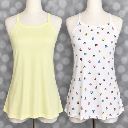 Two LuLaRoe Essential camis displayed on mannequin forms against a soft gray bokeh background featuring a solid butter yellow cami and a white cami with tiny multicolor retro floral print in pink, blue, yellow, and burgundy.